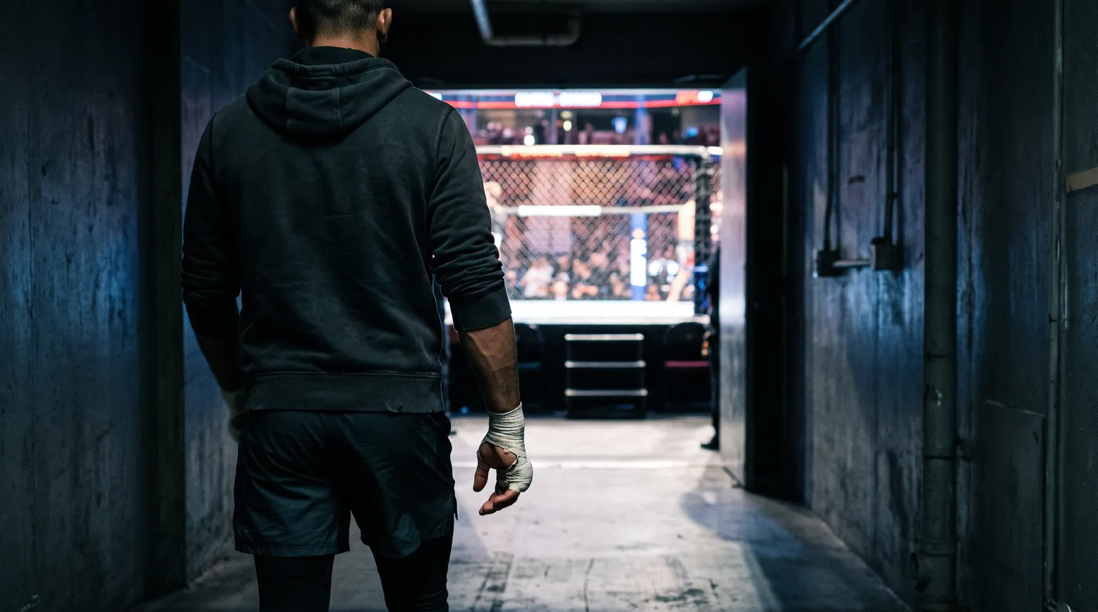 A fighter seen from behind walking down a dark hallway toward a brightly lit octagon